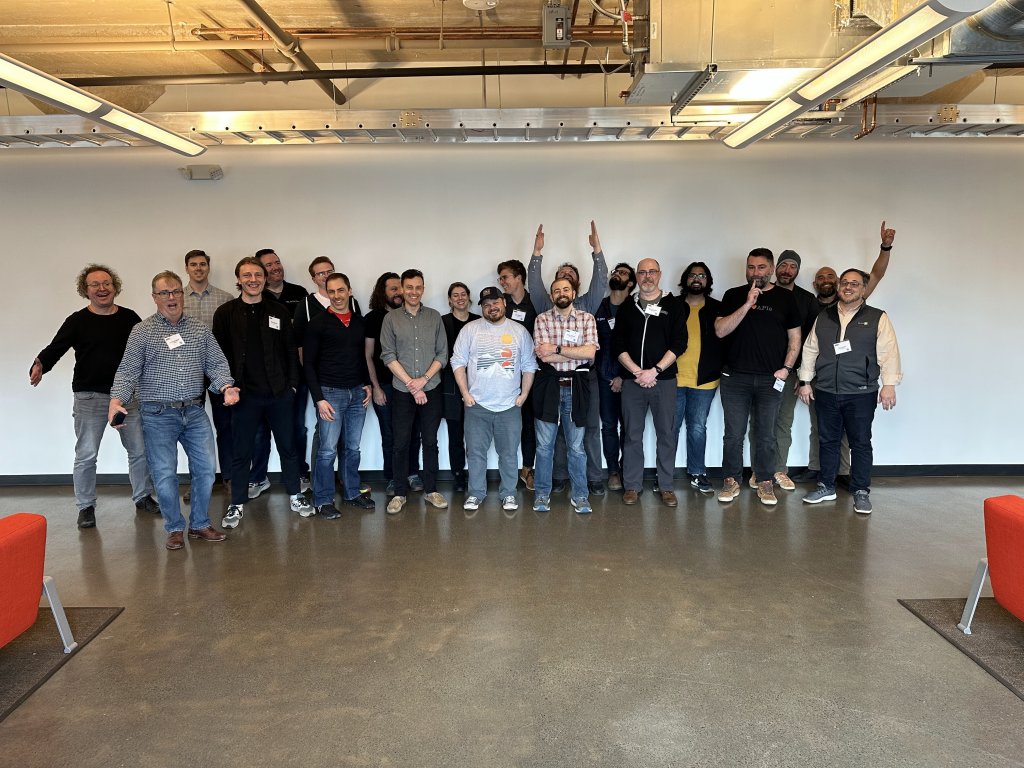 A group of approximately 20 attendees pose in front of a white wall making various silly expressions. 