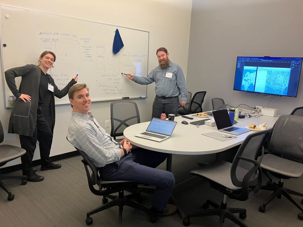 Two tasking sprint attendees stand in front of a whiteboard pointing at notes from the sprint. One attendee sits at a table in front of a laptop. 